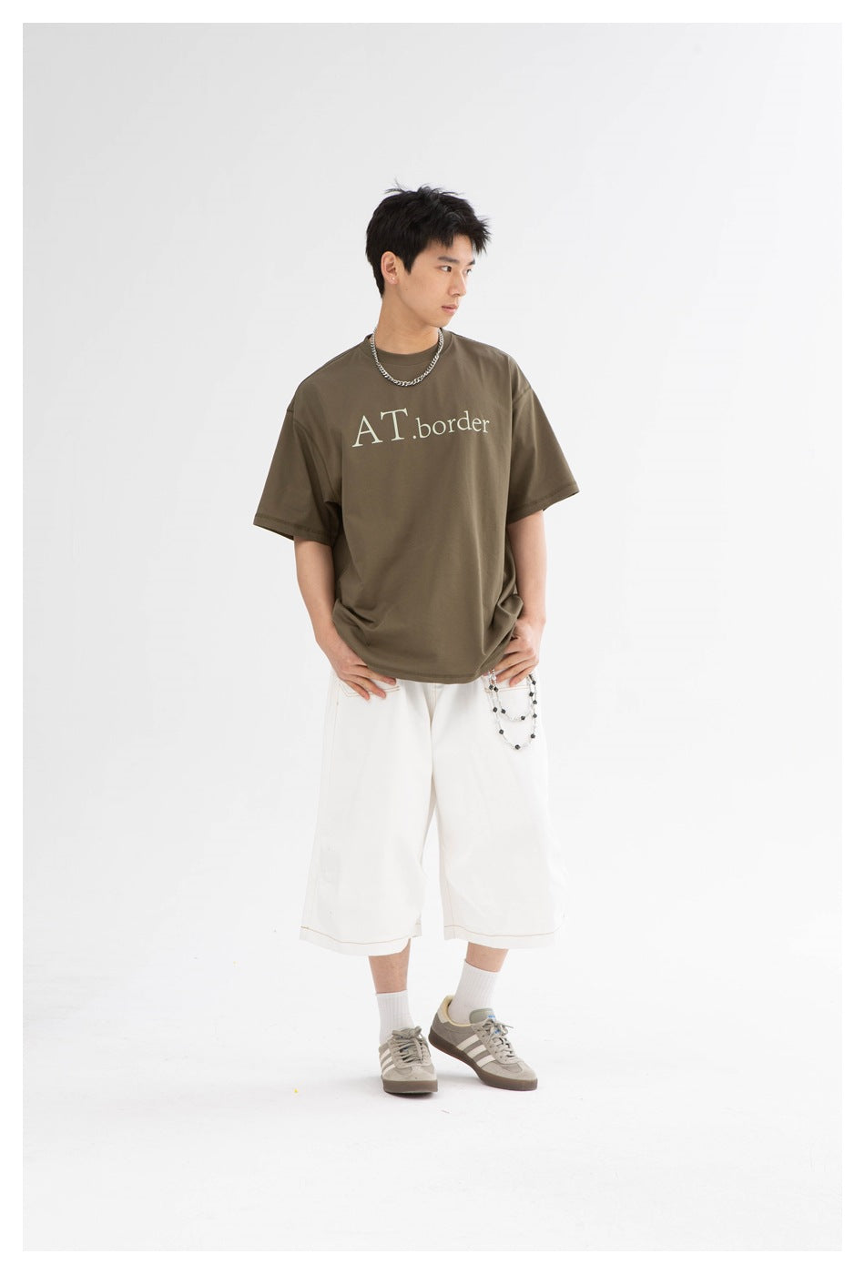 Model wearing an olive-brown graphic tee reading “AT.border,” white wide-leg shorts and light gray sneakers.