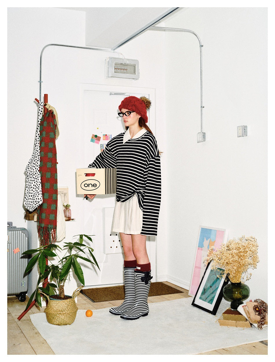 Model wearing a black-and-white striped oversized sweater with a white collar, cream pleated skirt, and red knit hat.