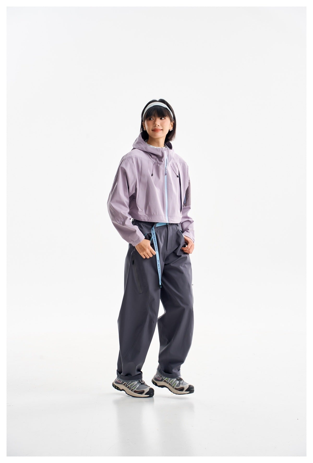 Model wearing a lavender cropped hooded jacket with dark, loose trousers and sneakers against a white background.