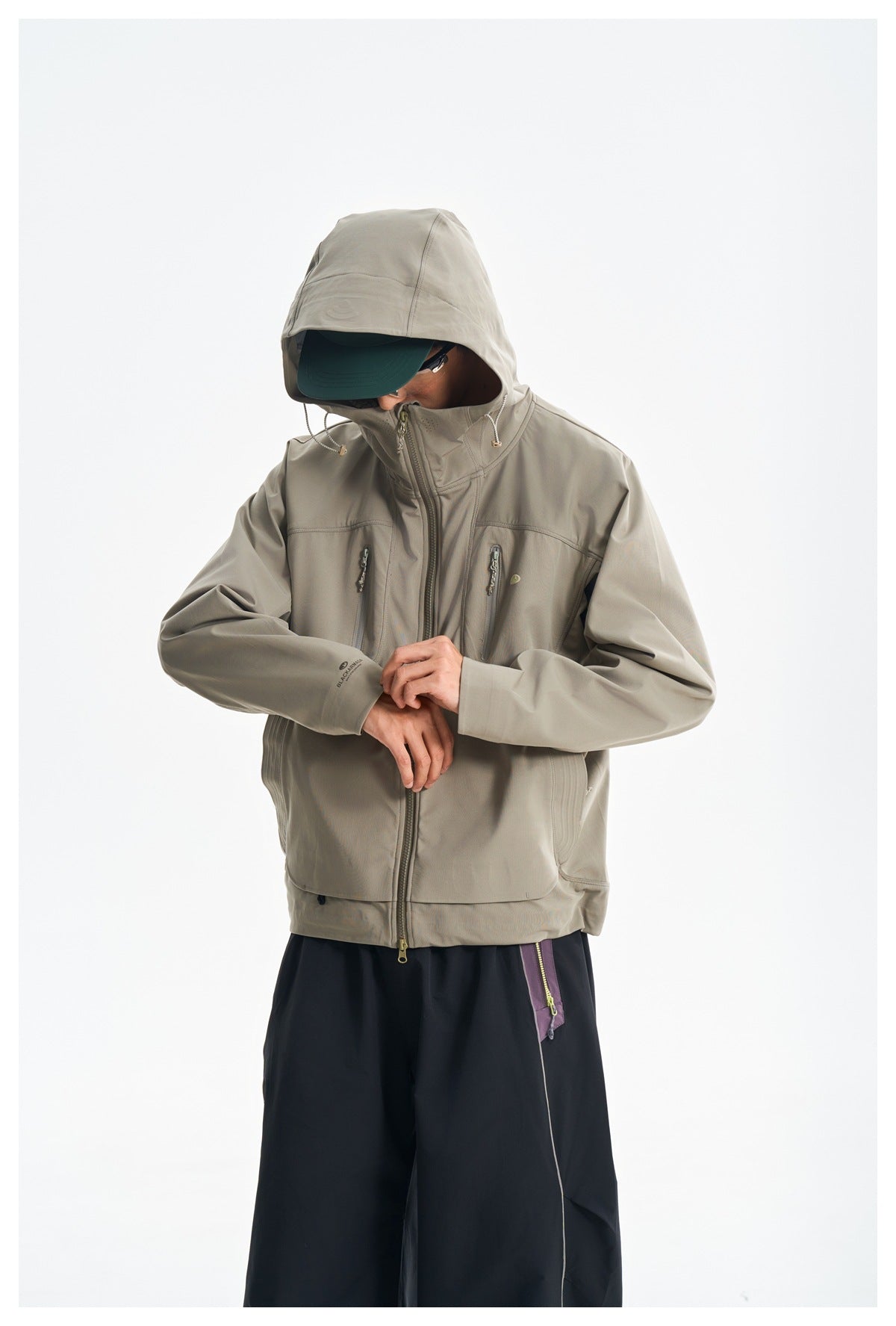 Model wearing a light gray-beige hooded jacket with two vertical chest zippers and dark pants.