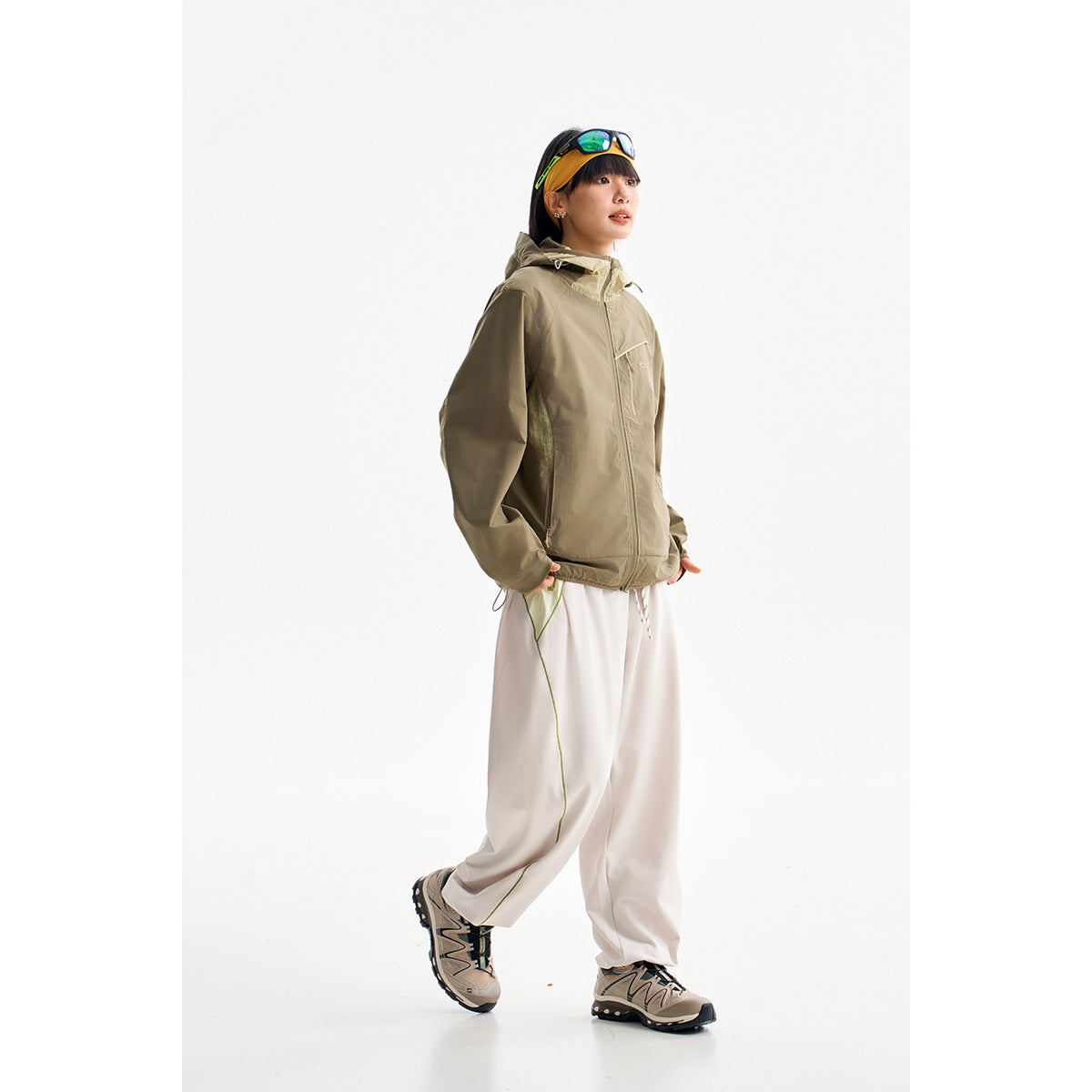 Model wearing an olive hooded zip jacket and loose white pants against a white studio background.