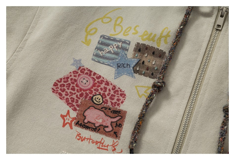 Close-up of colorful doodle-style prints on a beige hoodie beside its zipper and braided drawstring.