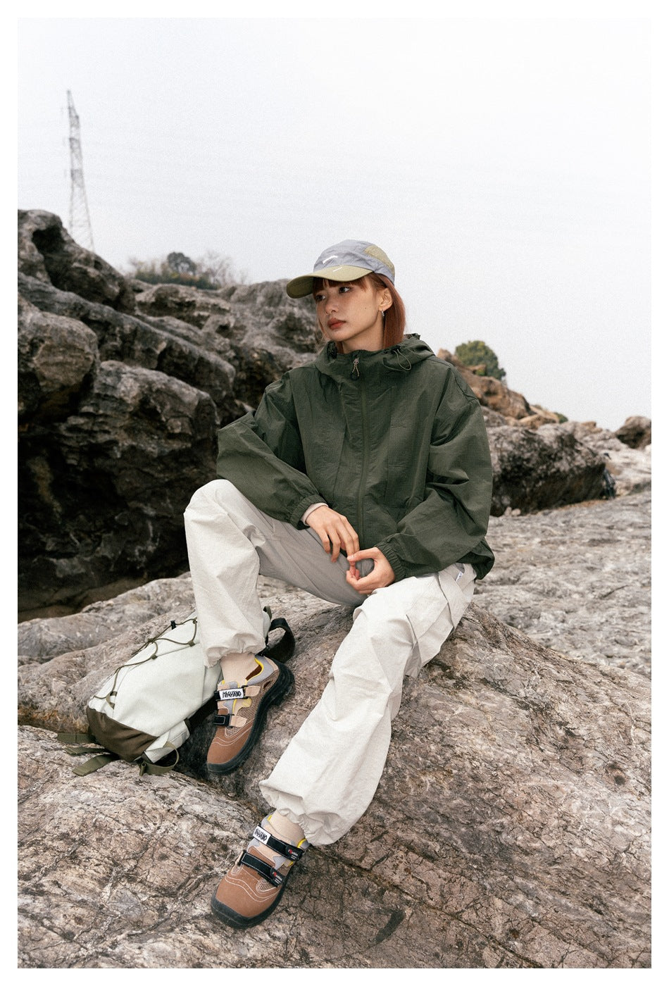 Model seated on rocks wearing a dark green hooded jacket, cream cargo pants, and a gray-and-beige cap.