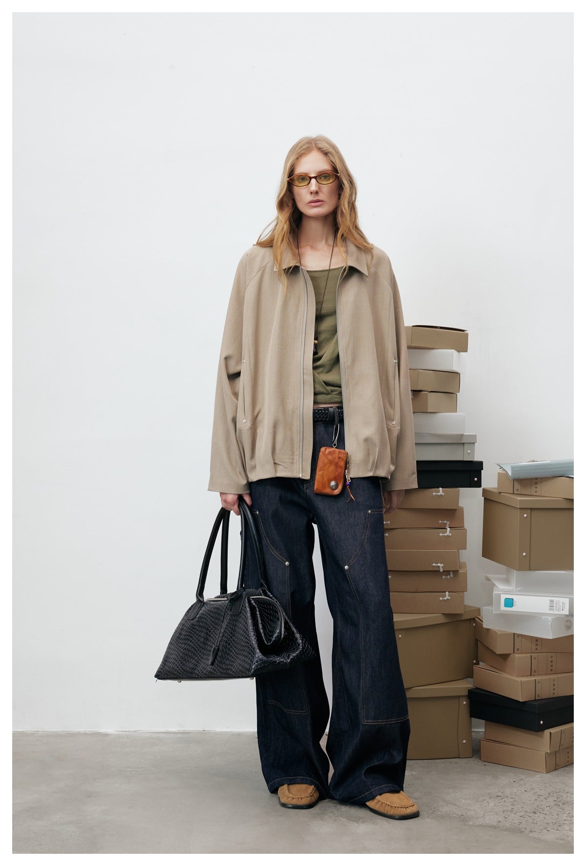 Model wearing an open khaki zip jacket over an olive top with wide dark denim jeans, standing beside stacked boxes