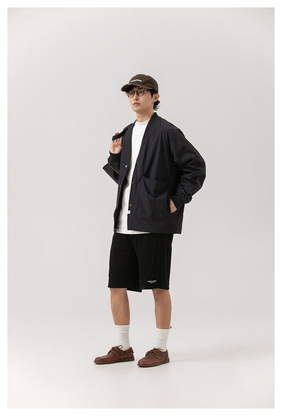 Full-length model wearing a black snap-front jacket over a white shirt, paired with black shorts and brown shoes.