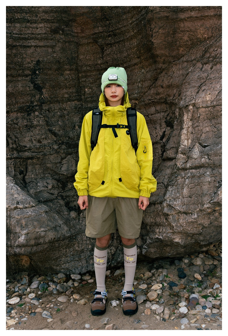 Model wearing a bright yellow hooded jacket with a front zip, large pockets, and taupe shorts.