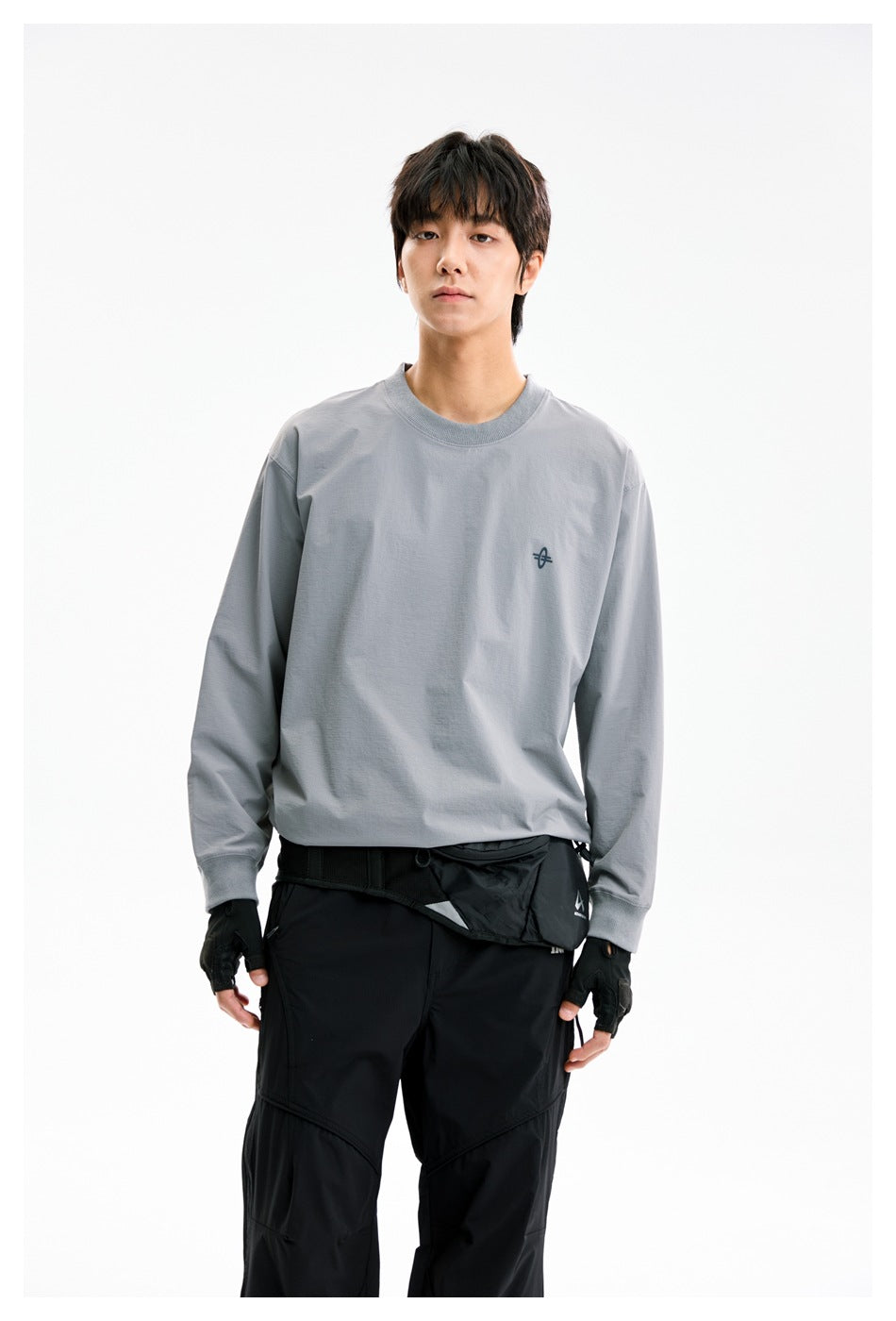 Front view of a model wearing a gray long-sleeve crewneck with a small chest emblem and black pants.