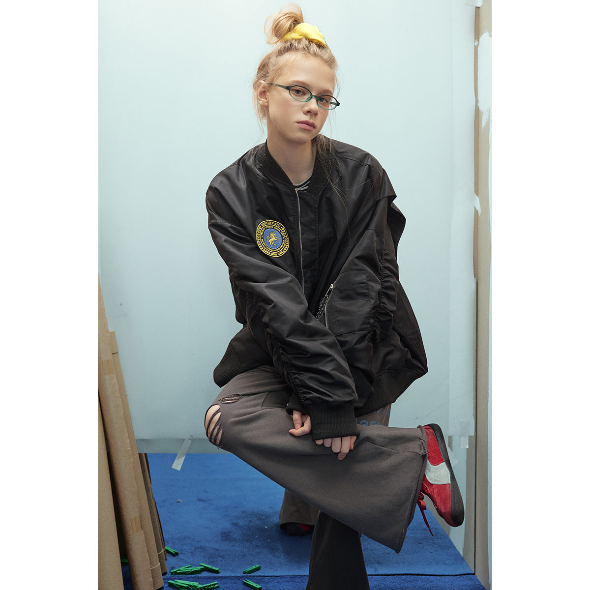Model in an oversized black bomber jacket with a chest patch and sleeve pocket, seated in dark trousers.