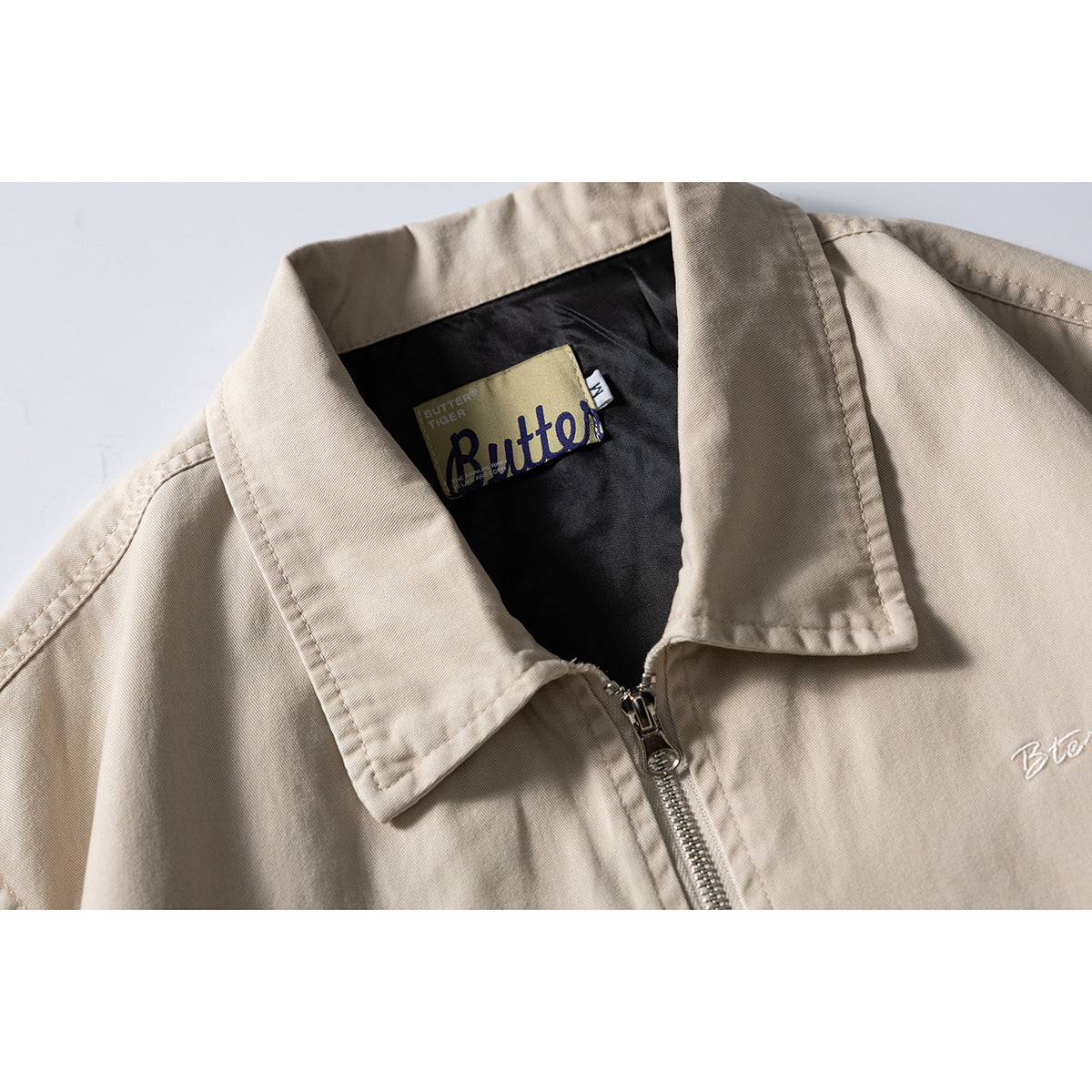 Close-up of a beige zip-front jacket collar, inner label, zipper and white chest embroidery.