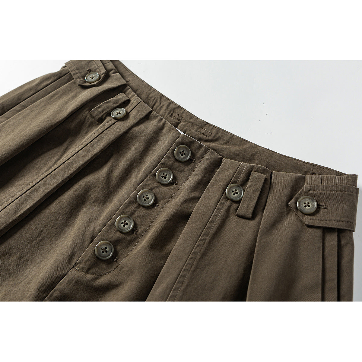 Close-up of brown trousers showing the high waist, pleats, and a row of front buttons.