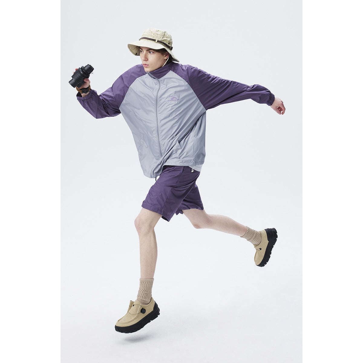 Model leaping in a light gray jacket with purple sleeves and matching shorts, holding binoculars.
