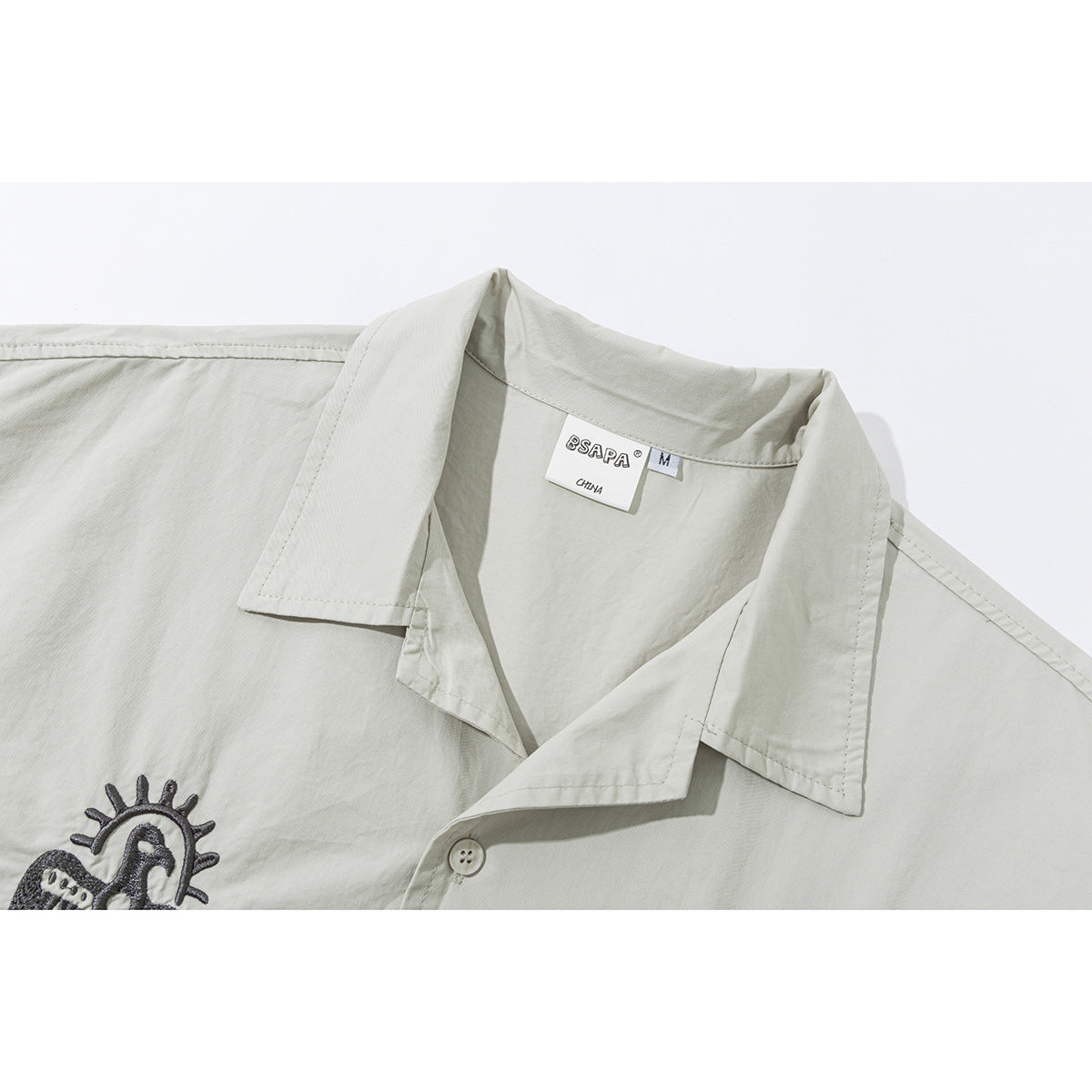 Close-up of a pale gray shirt’s pointed collar, button, size tag, and part of a dark embroidered motif.