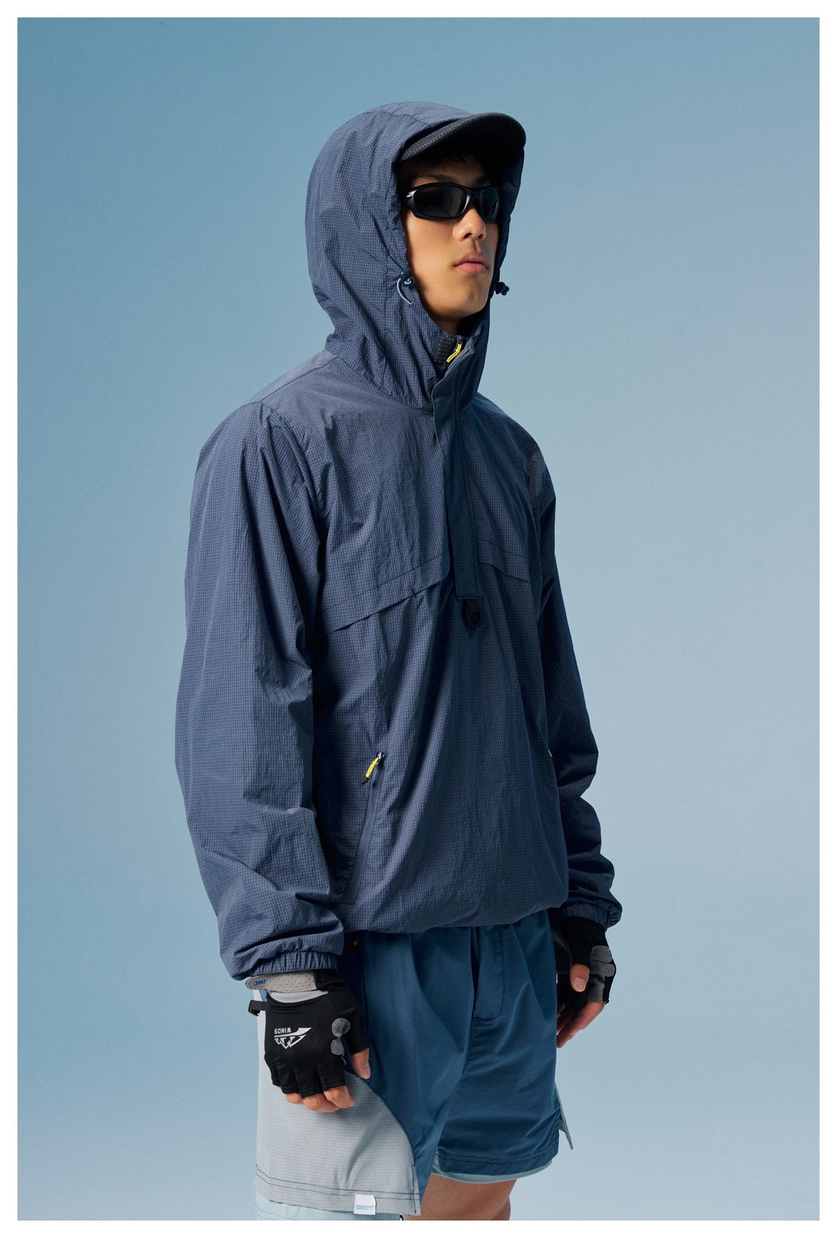 Model wearing a slate-blue grid-pattern hooded anorak with yellow zipper pulls and dark blue shorts.