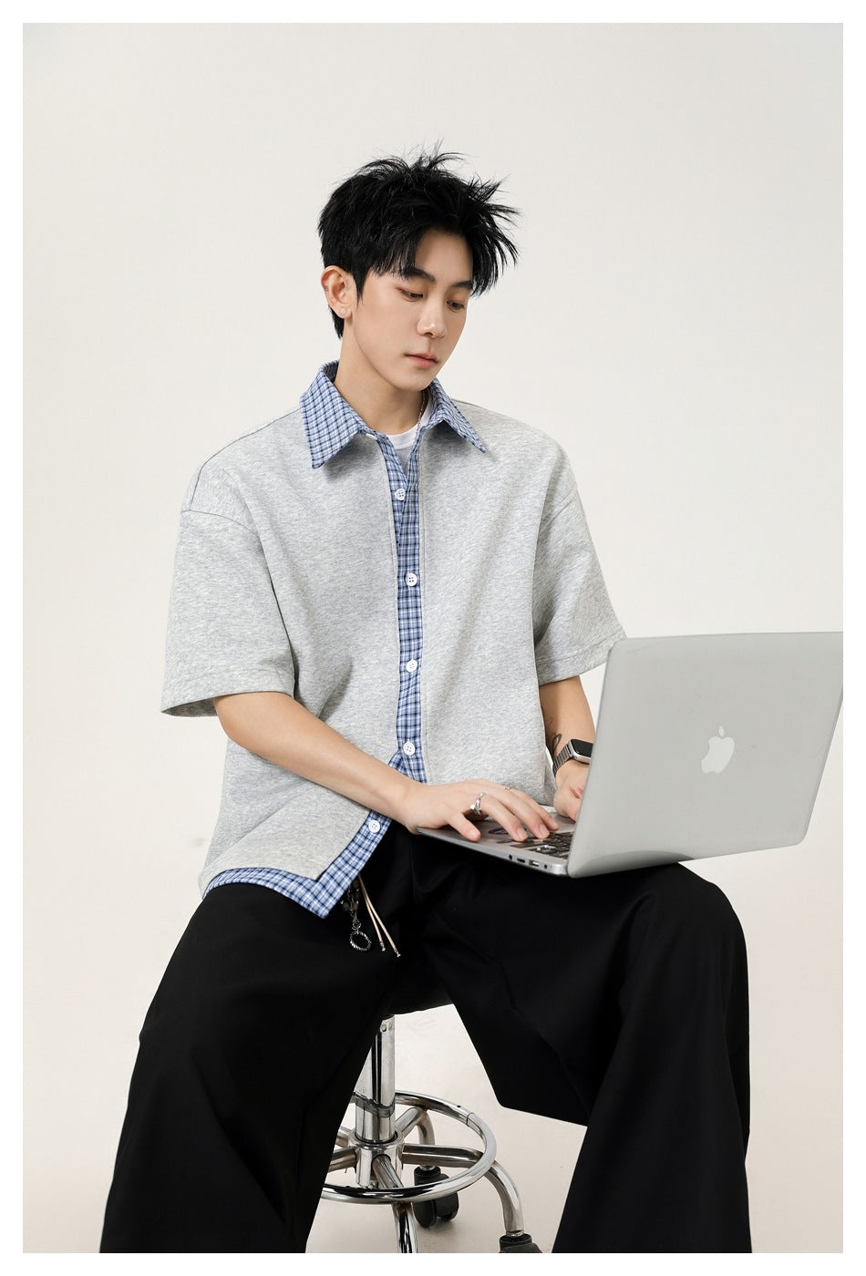 Man seated on a stool using a laptop, wearing a gray short-sleeve top with blue plaid collar and placket and black pants