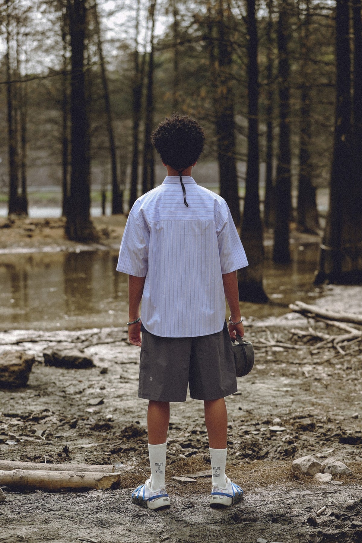 Back view of a person in a light blue striped short-sleeve shirt, gray shorts and white socks by a forest pond