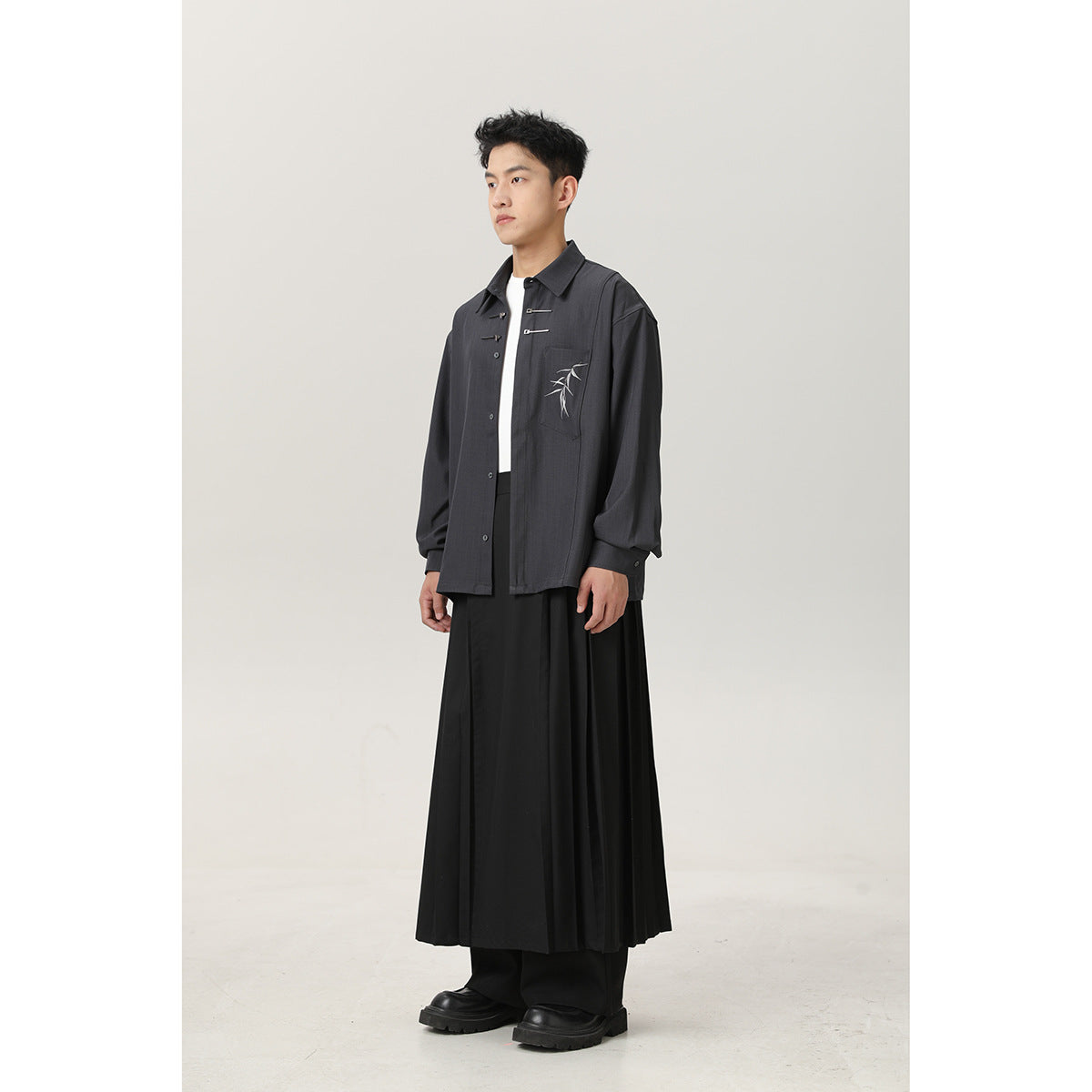 Full-length view of a man wearing an open charcoal shirt over a white top, a long black pleated skirt over trousers, and