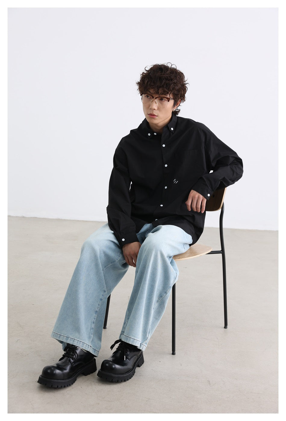 Seated model wearing a loose black button-down shirt with a chest pocket and white buttons.