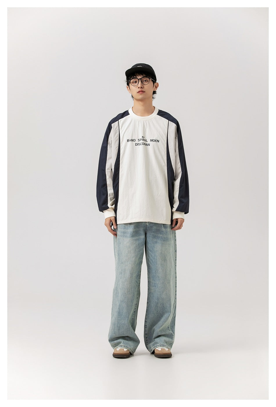 Model wearing cream and navy color-block sweatshirt with light-wash wide-leg jeans and black cap, front view