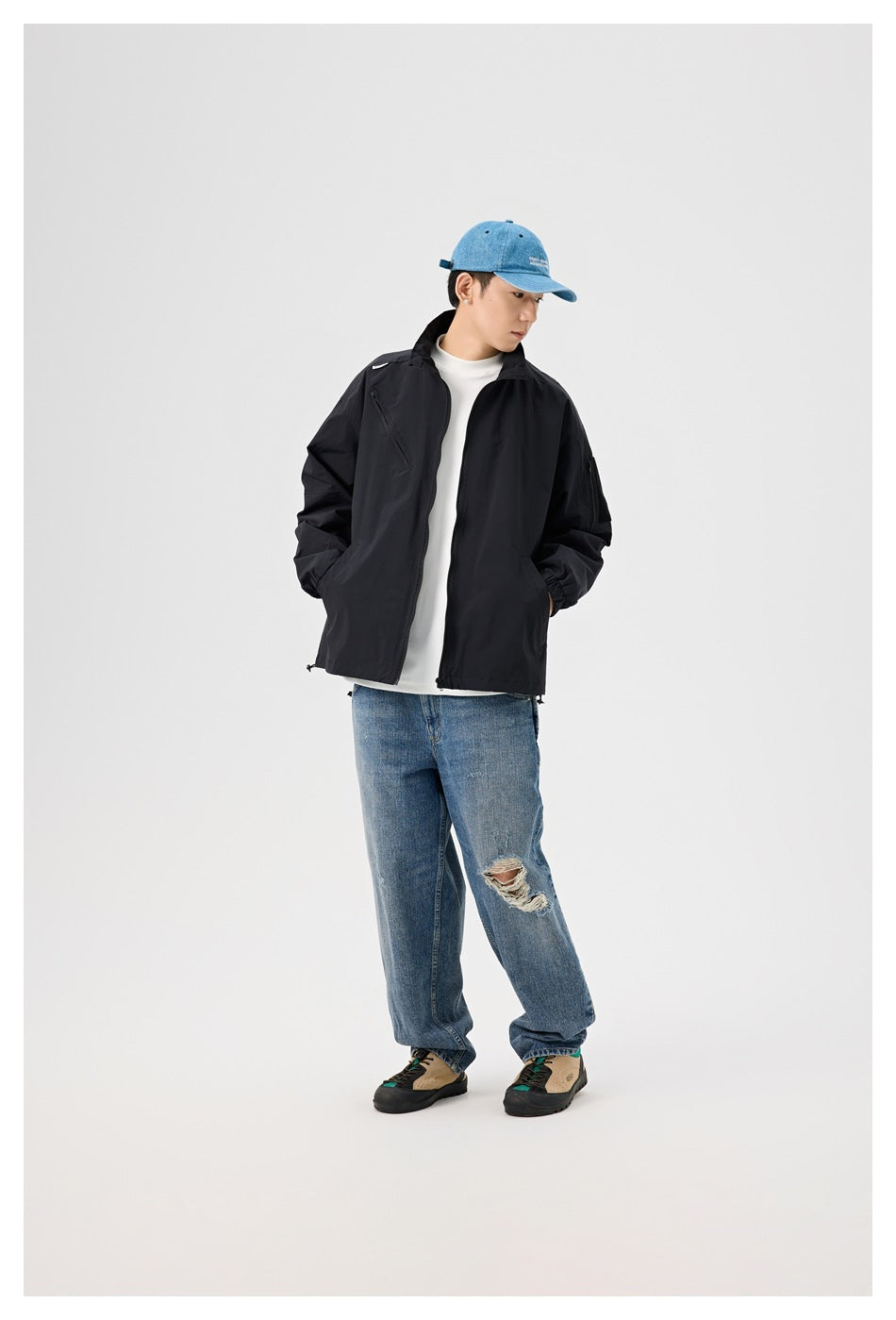 Model wearing a black zip jacket over a white tee with blue jeans, denim cap and sneakers, full-length view