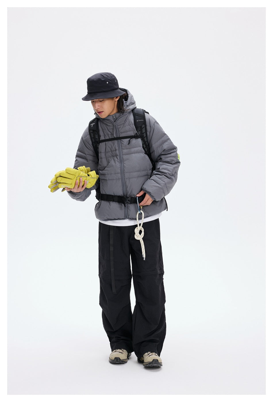 Model wearing a gray hooded puffer jacket with a backpack, bucket hat, black pants and yellow gloves