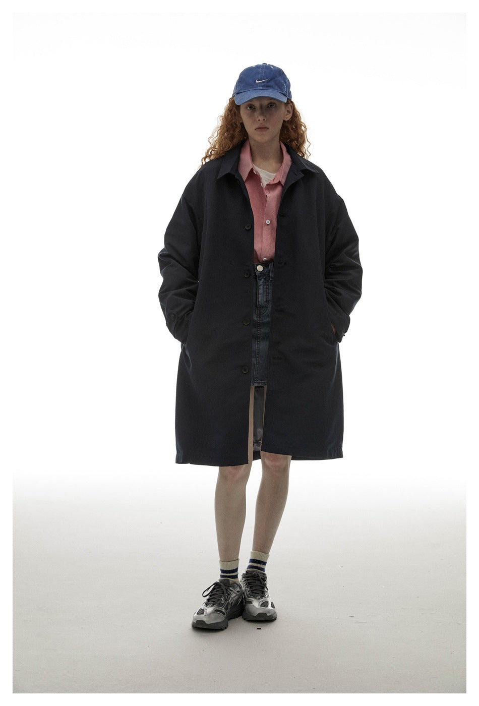 Model wearing the navy button-front coat open over a pink shirt and denim skirt, with a blue cap and sneakers