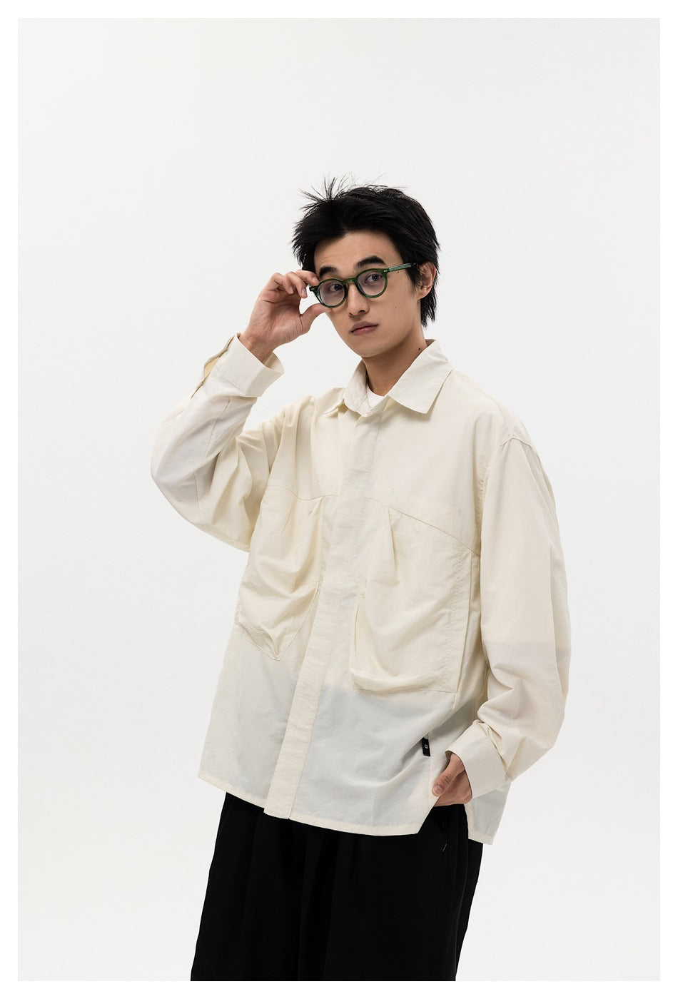 Model wearing an off-white oversized pocket shirt with green glasses and black trousers, one hand raised to glasses