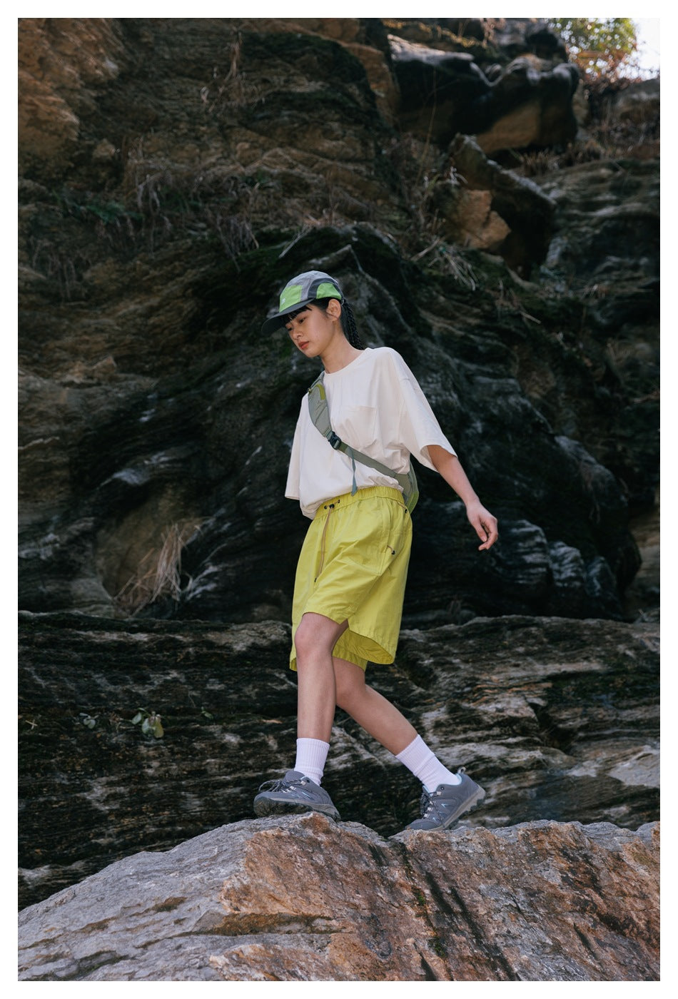 Model wearing a white pocket T-shirt and bright yellow hiking shorts on rocky terrain.