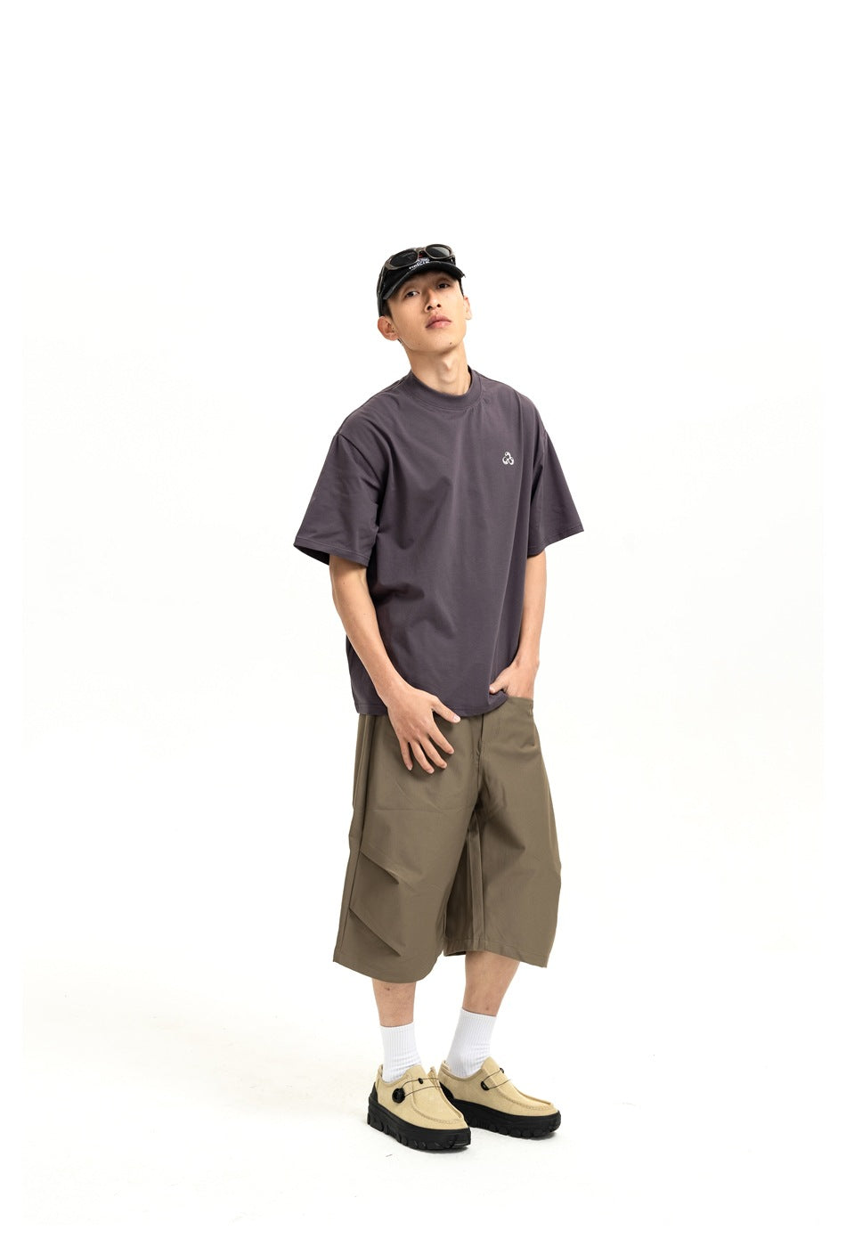 Model posing in khaki-brown wide-leg shorts with angled pleat detail, gray T-shirt, white socks and beige shoes