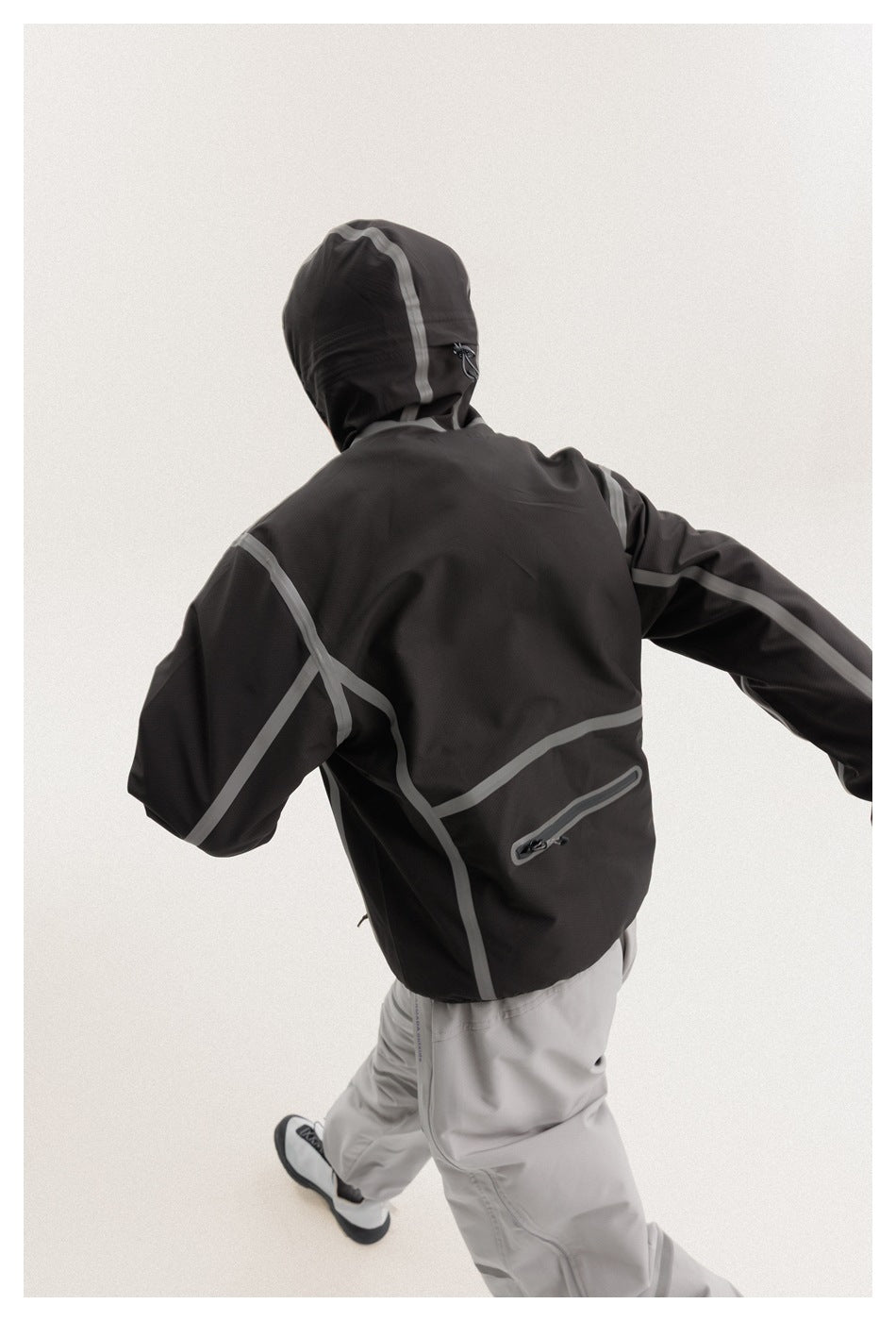 Back view of a model in a black hooded jacket with gray panel piping and a horizontal back zipper pocket.