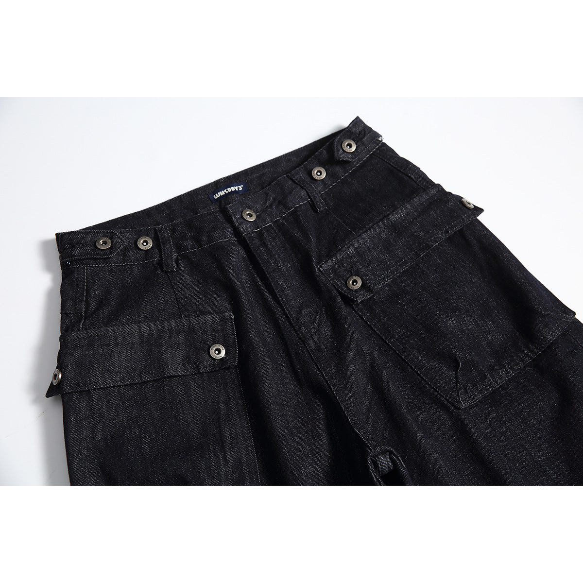 Close-up of black denim trousers with buttoned waistband tabs and large front utility pockets.