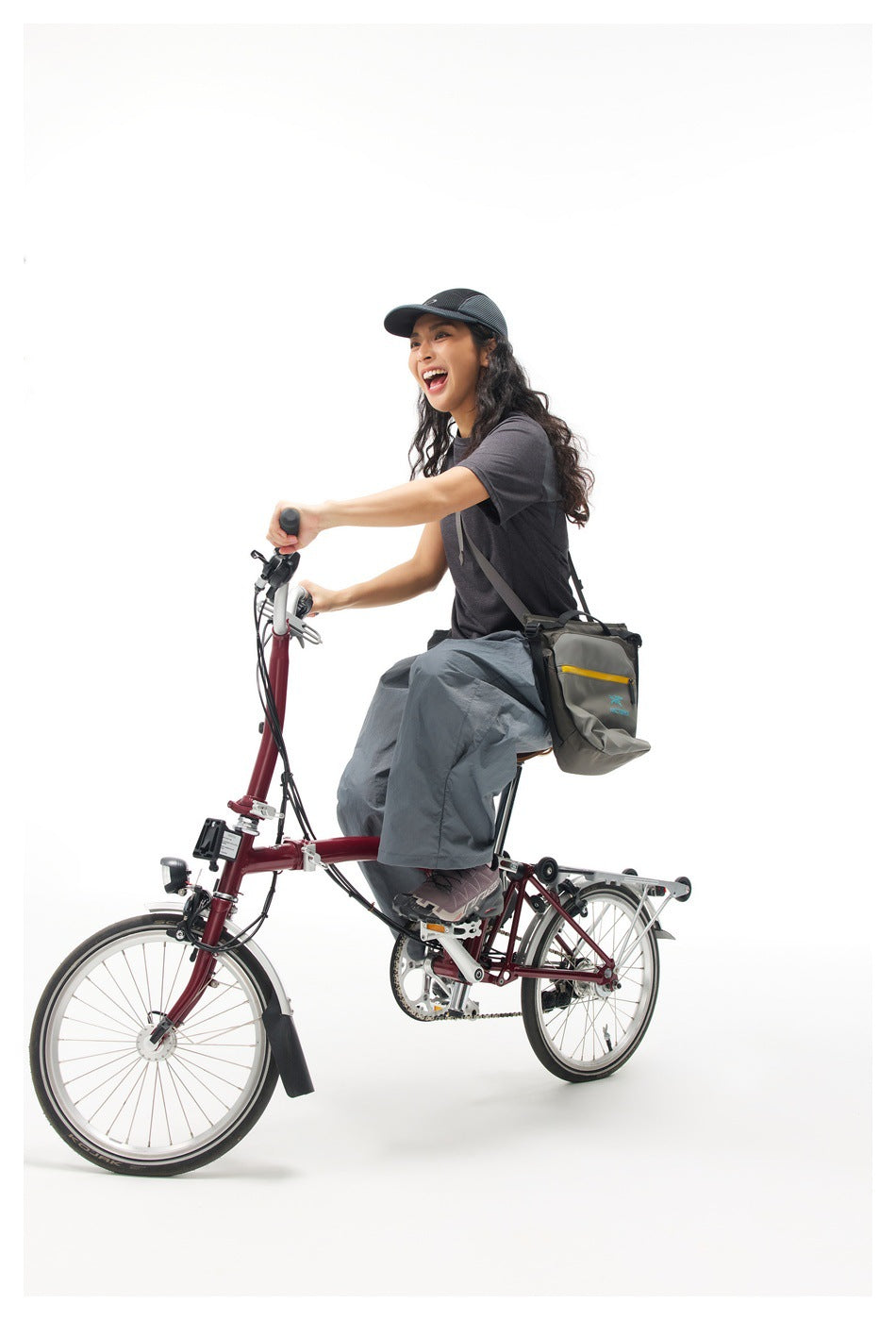 Model riding a bicycle in a charcoal T-shirt, loose slate-gray pants, and a black cap, with a shoulder bag.