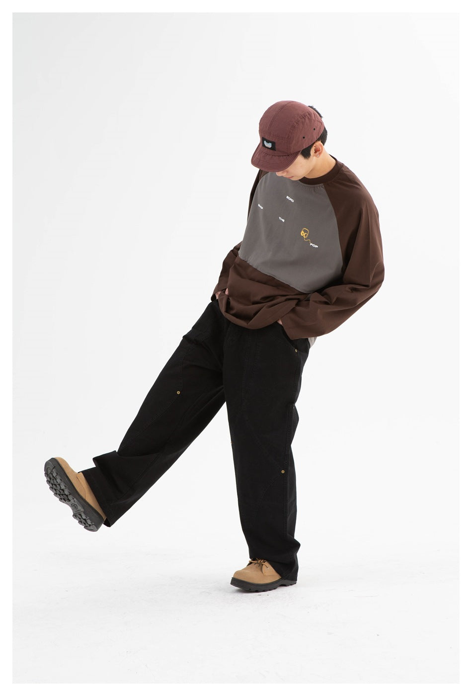 Model in a brown and gray color-block long-sleeve shirt, black pants, and burgundy cap, leaning with one leg raised.