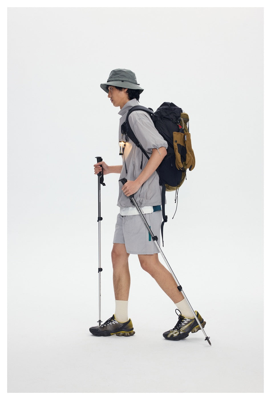 Side view of a model wearing a light gray jacket and shorts with a bucket hat and hiking backpack.