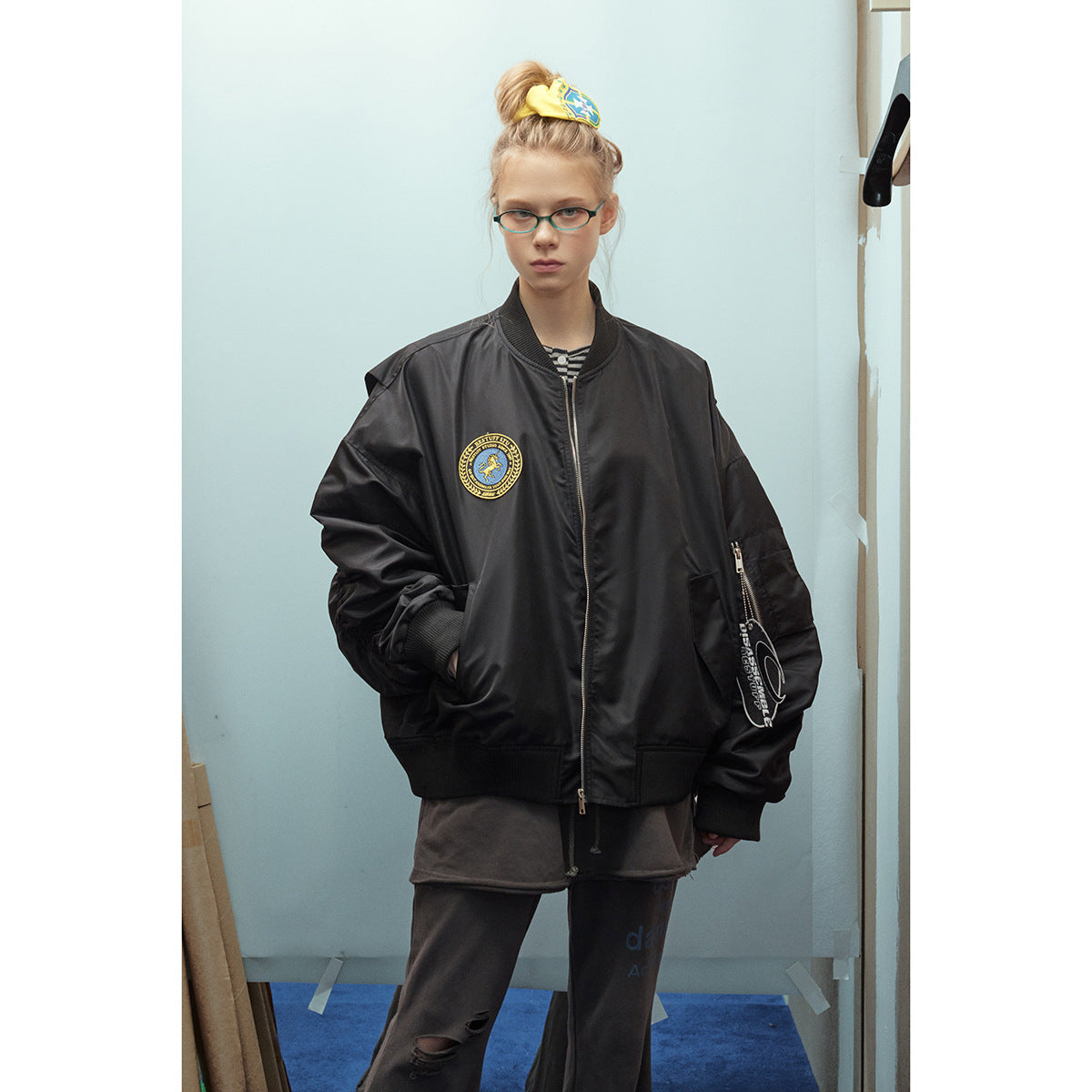 Model wearing an oversized black zip-front bomber jacket with a chest patch and sleeve graphics, over dark clothes.