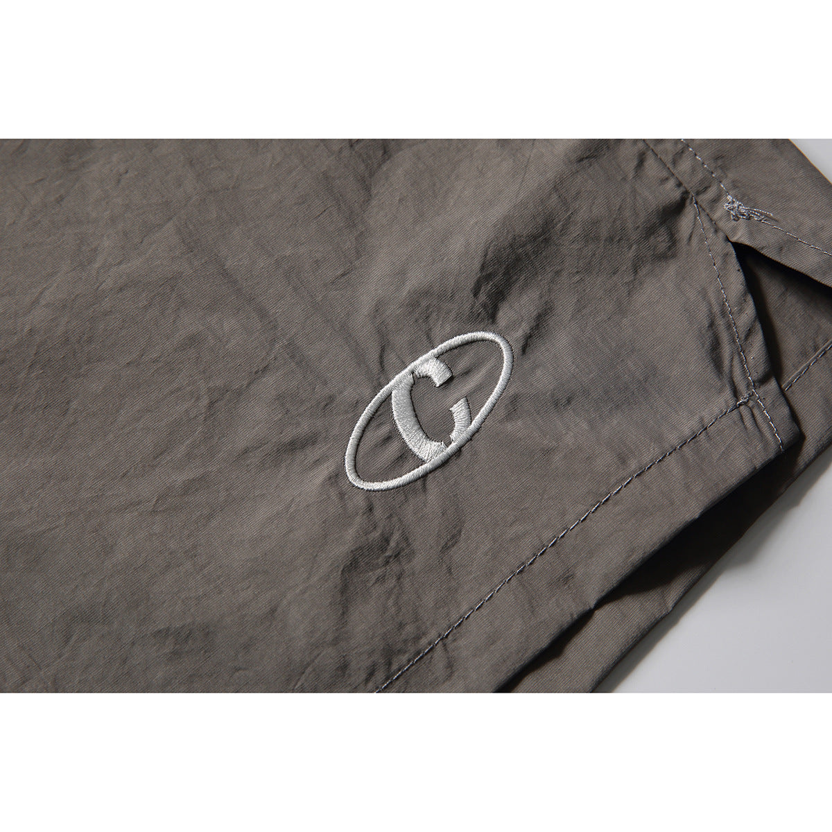 Close-up of gray shorts with a small white embroidered oval emblem near the hem.