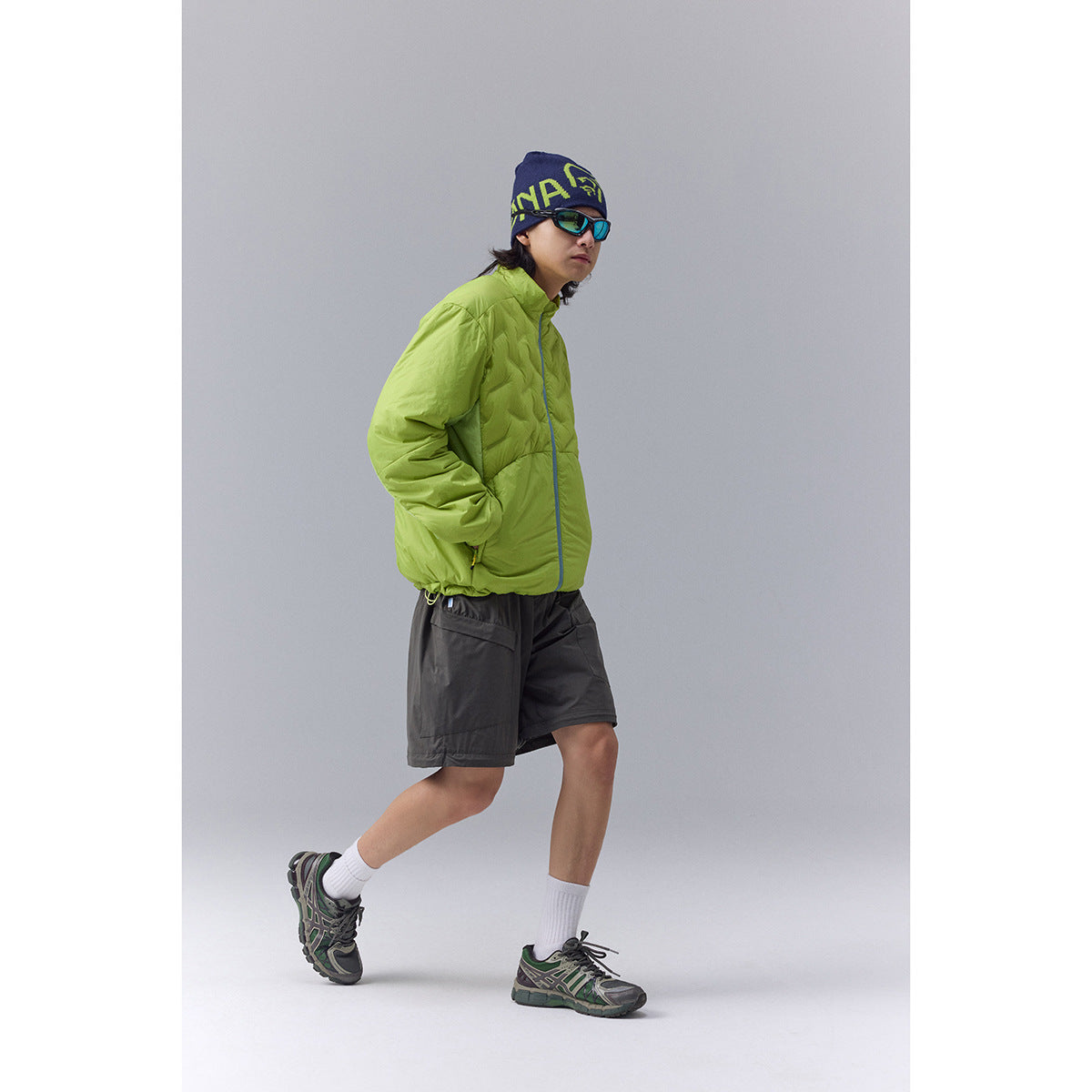 Model wears a bright lime quilted jacket with blue trim, charcoal shorts, a graphic beanie, sunglasses, and trail shoes.