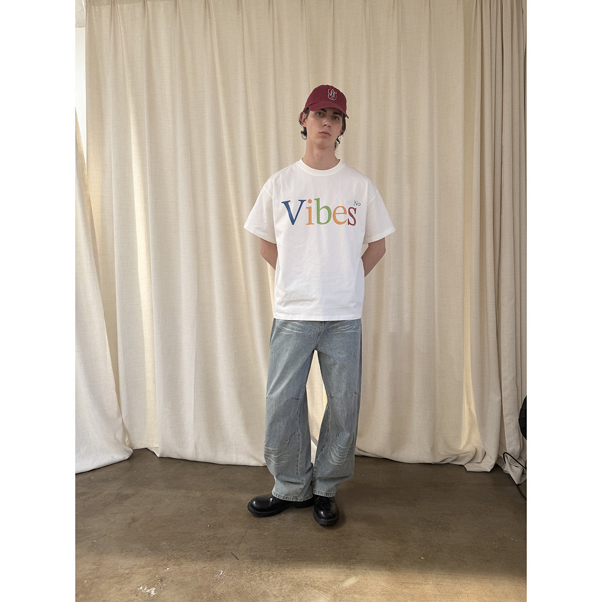 Model in a white T-shirt with multicolored “Vibes No” lettering, loose light-wash jeans and a burgundy cap.