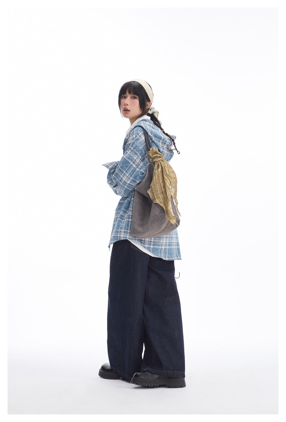 Model in a blue-and-white plaid hooded shirt jacket and wide dark jeans, shown at an angle with a shoulder bag.