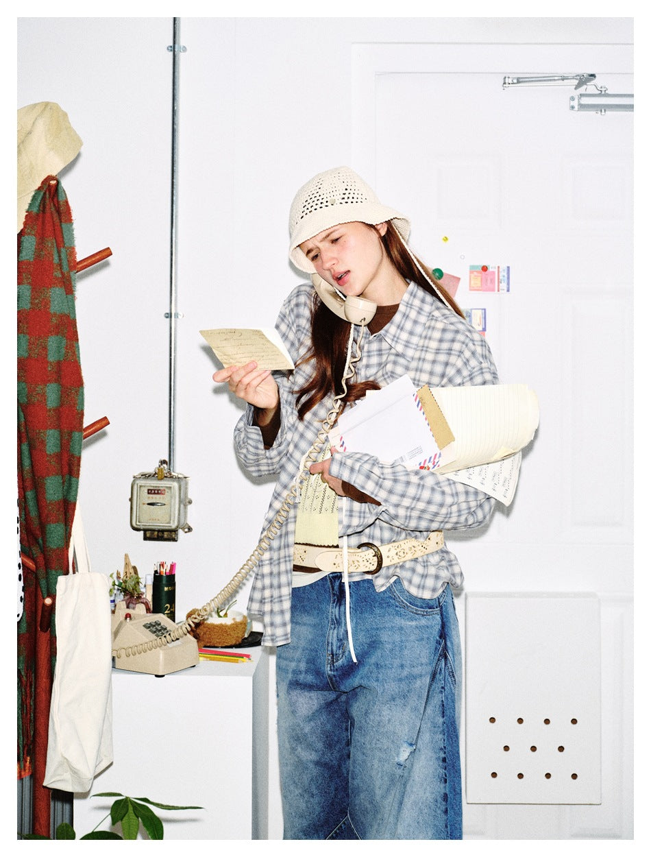 Model in a blue-gray plaid button-up shirt and cream crochet hat, holding papers beside a telephone.