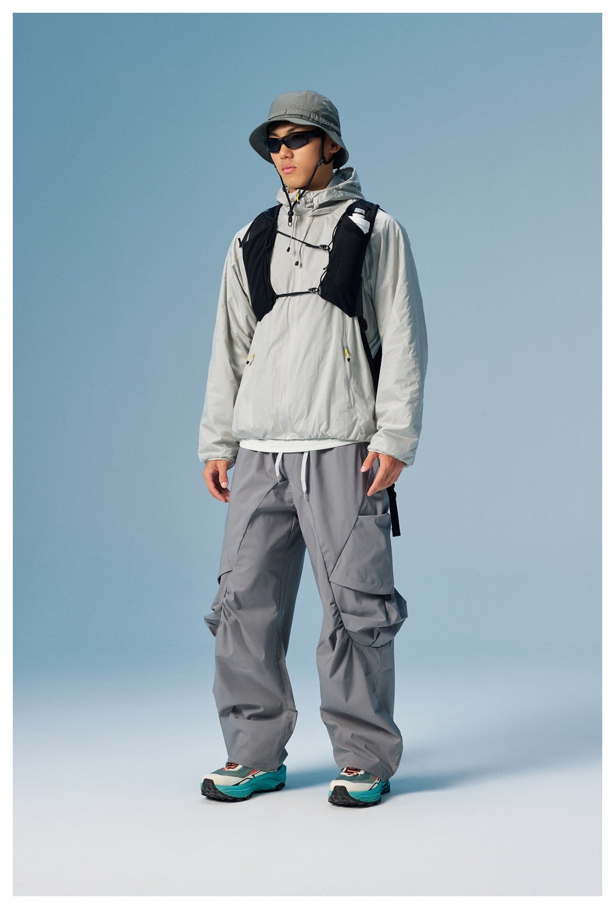 Model wearing a light gray hooded windbreaker and gray cargo pants with a black running vest and bucket hat.