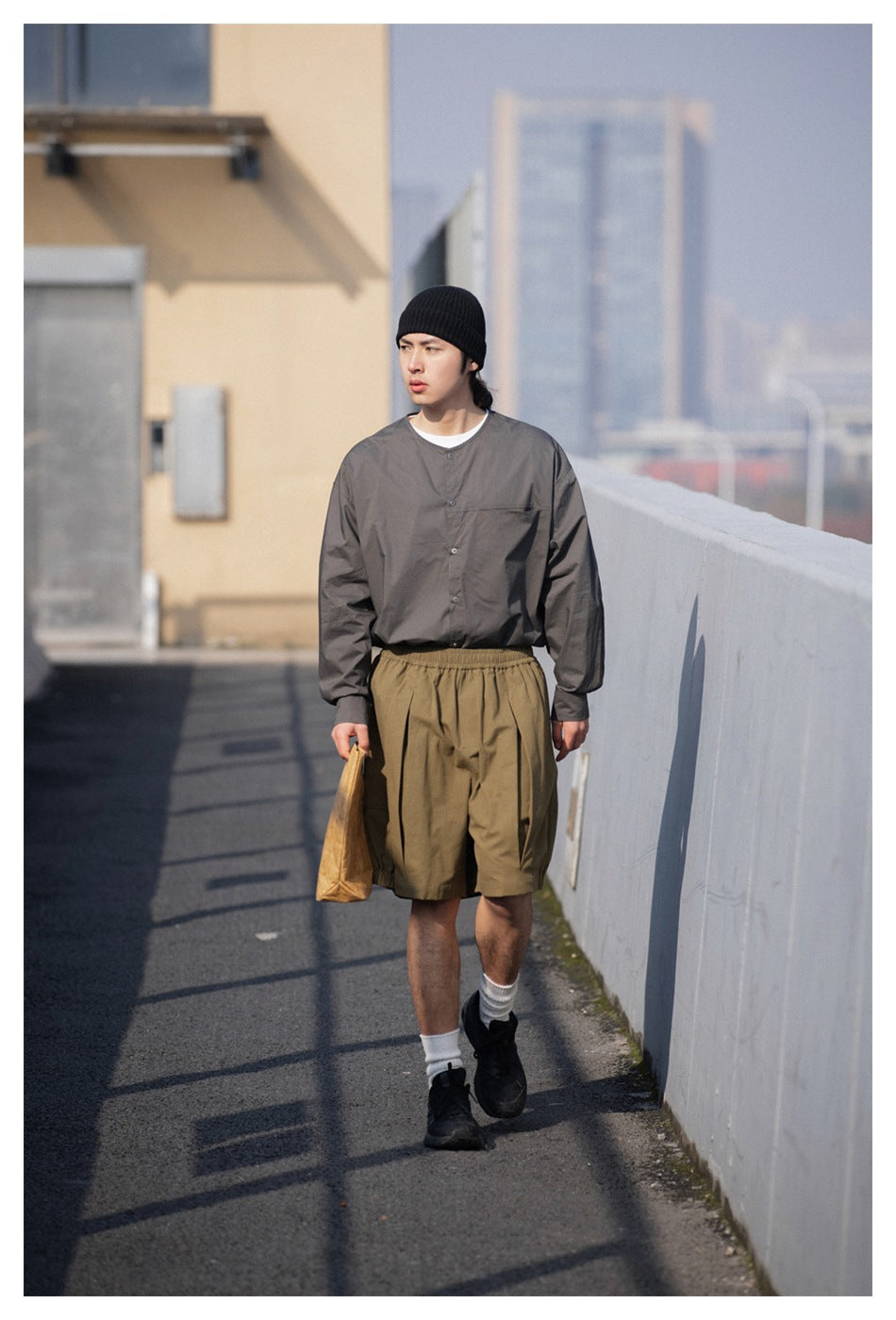 Man walking outdoors in olive pleated knee-length shorts with a gray collarless shirt, black beanie, and sneakers