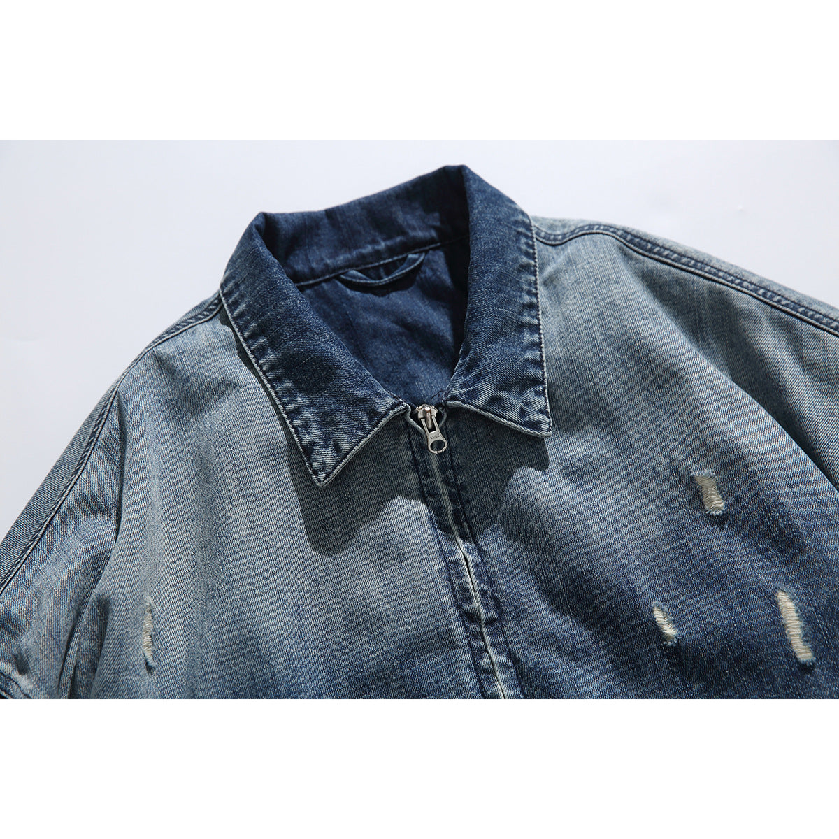 Close-up of a blue distressed denim jacket showing its pointed collar, front zipper and small frayed tears.