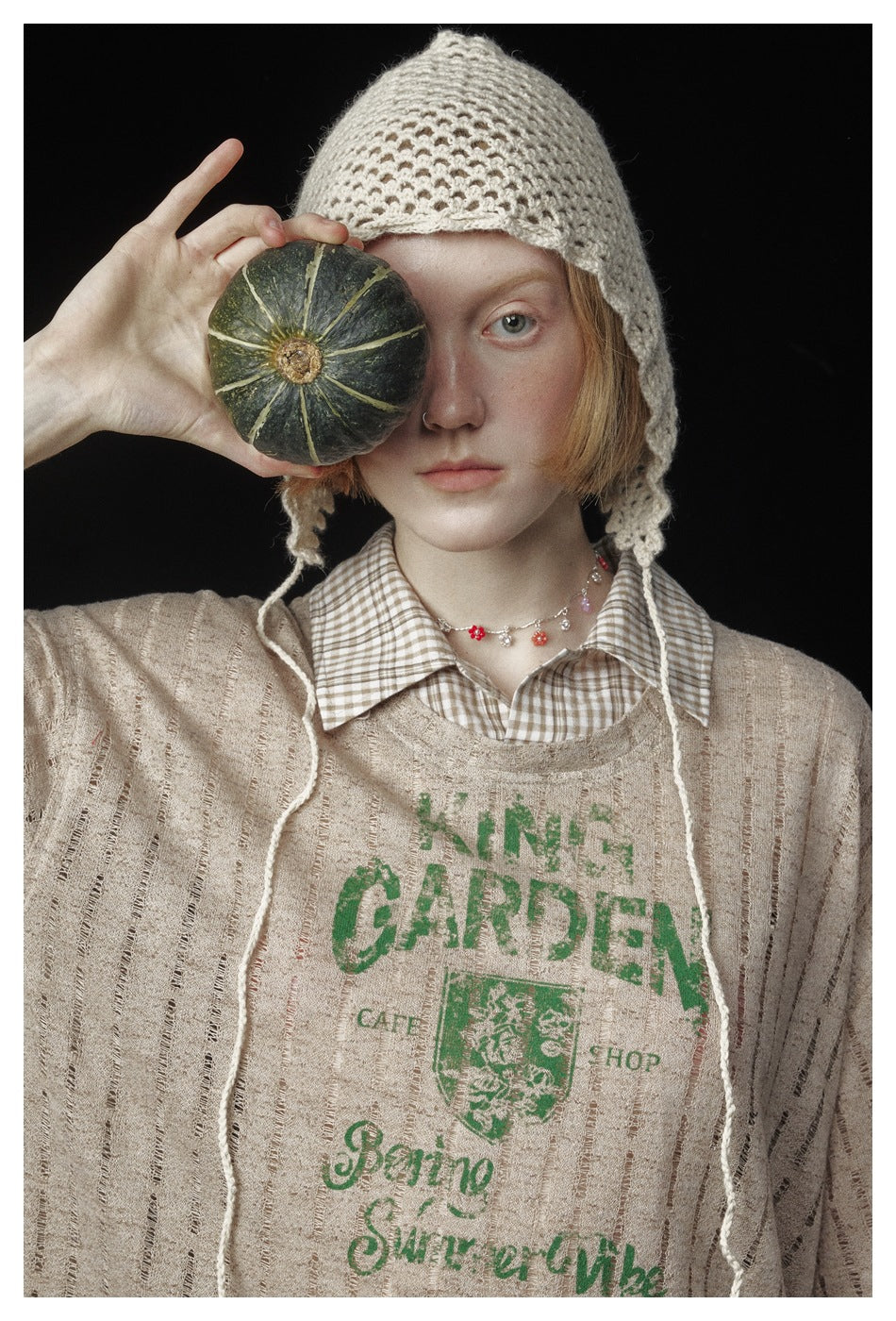 Close portrait of a model in a beige striped graphic top, checked collar and cream crocheted bonnet, holding a pumpkin over