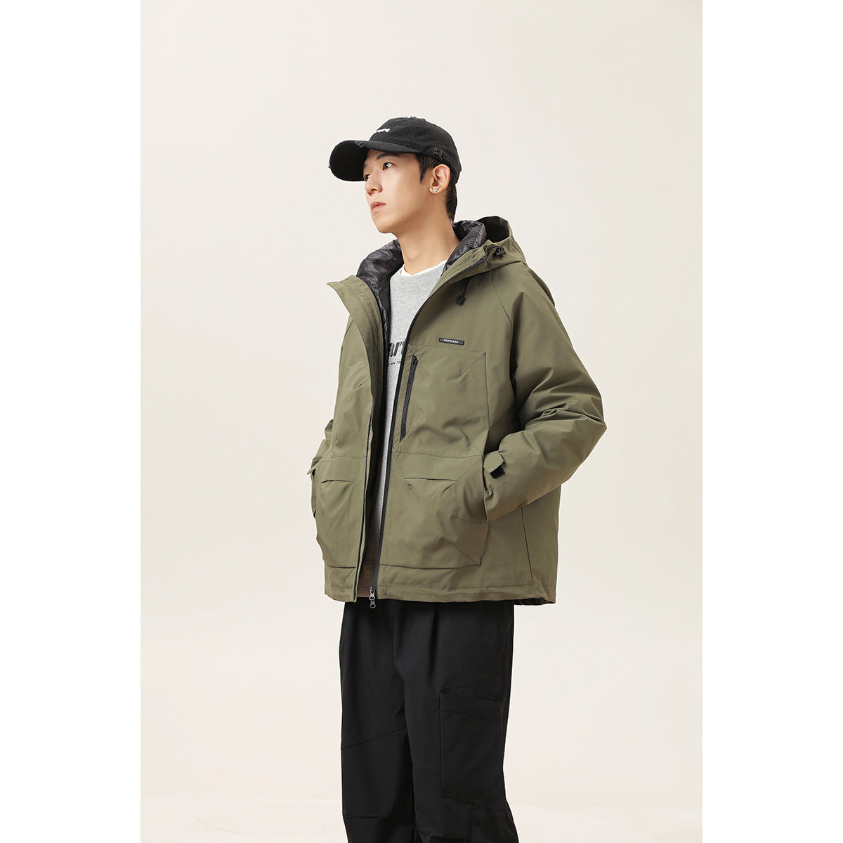 Model wearing an olive hooded jacket with front pockets and a black cap, shown from the front at an angle.