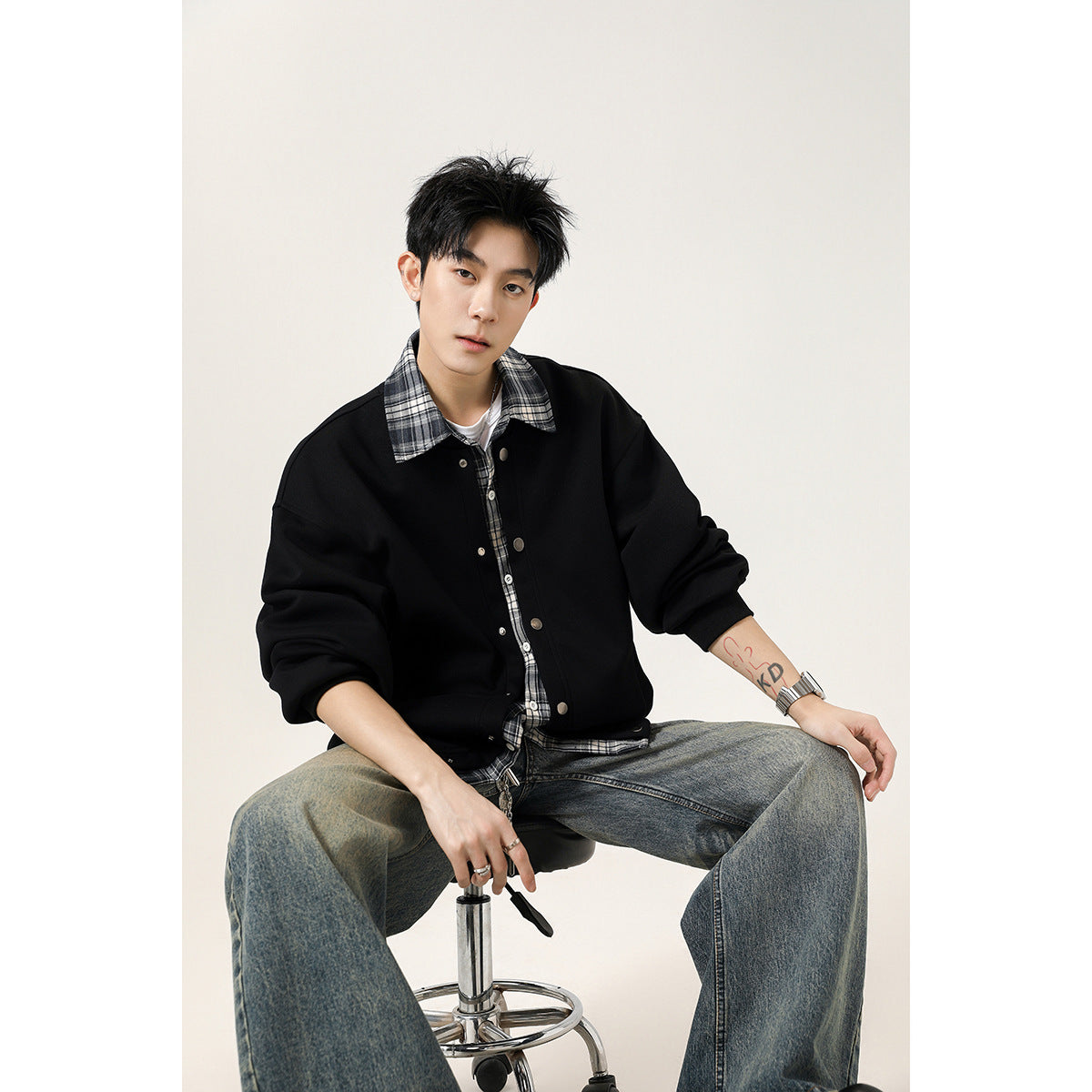 Seated model wearing a black snap-front jacket over a black-and-white plaid shirt.