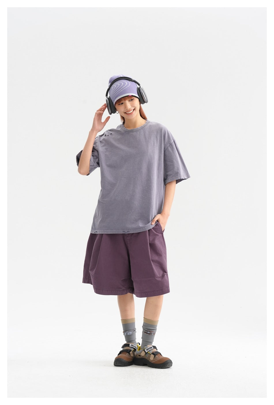 Model wearing an oversized faded lavender-gray T-shirt with small shoulder distressing and plum shorts, shown full length.