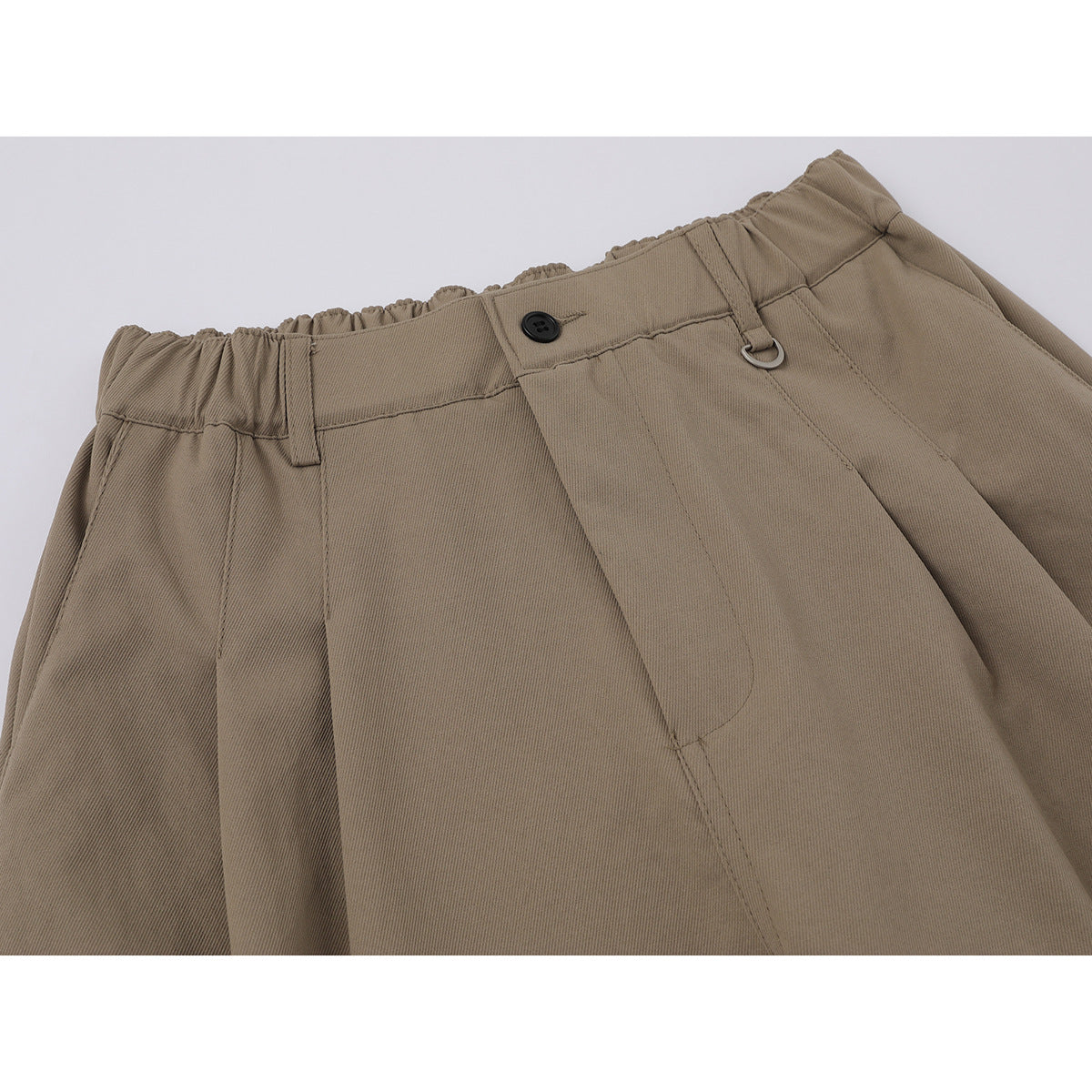 Close-up of taupe trousers showing the gathered waistband, button, belt loop, metal ring, and front pleats.