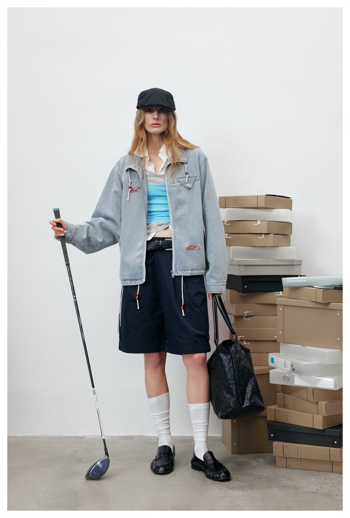 Model wearing the light-blue striped zip jacket open over a blue tank top and navy shorts, holding a golf club and bag
