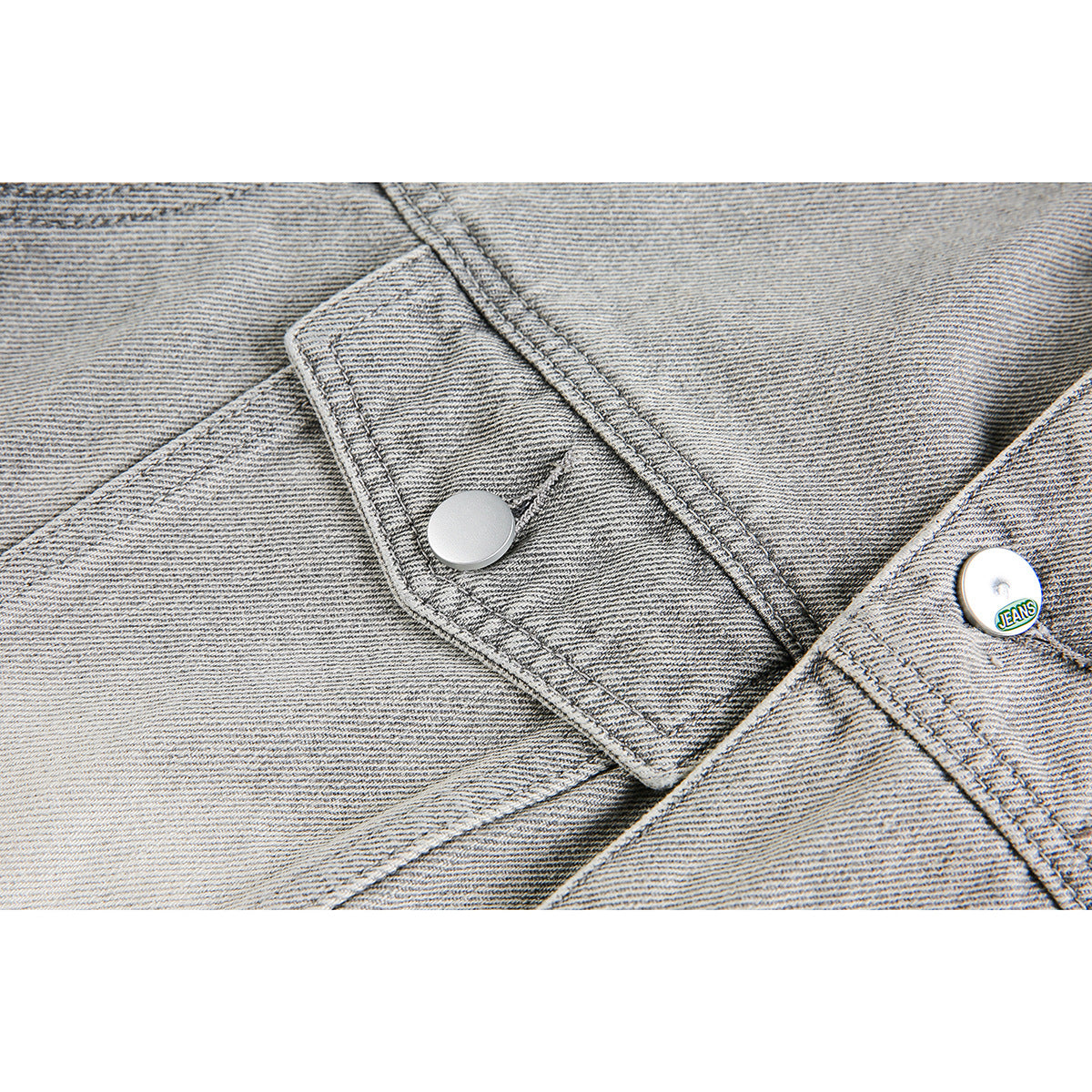 Close-up of a light gray jacket’s snap-button placket; one button has a small “JEANS” marking.