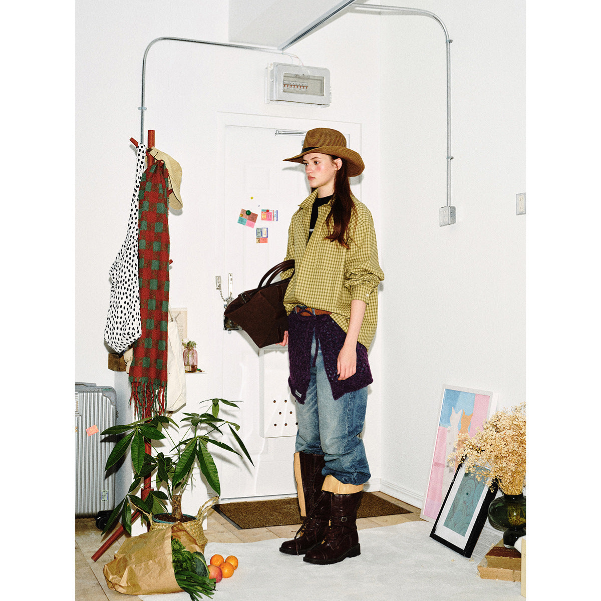 Model wearing a yellow check button-up shirt, jeans, dark boots and a brown hat in a room.